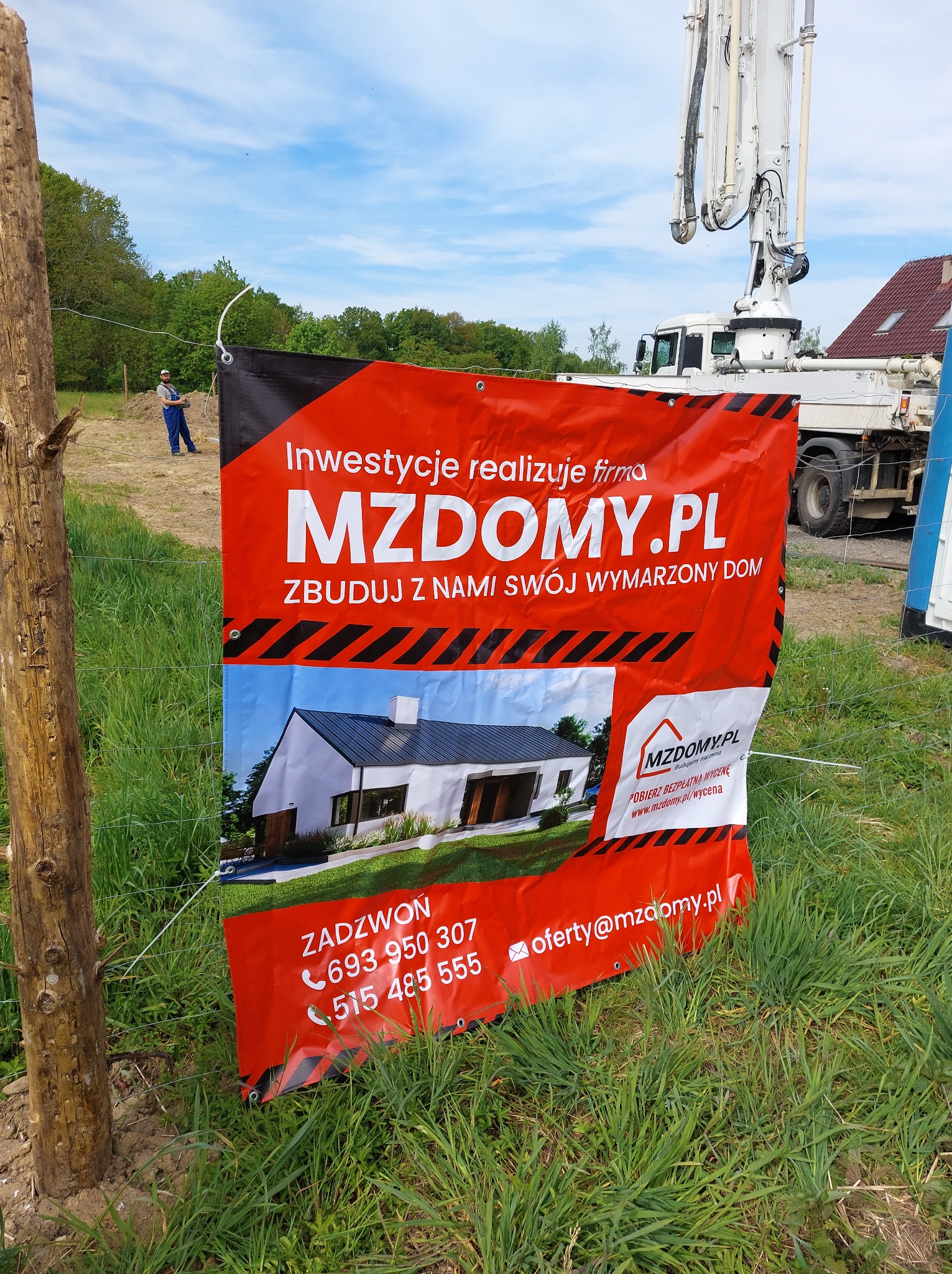Baner reklamowy firmy MZDOMY.PL na tle placu budowy z widoczną pompą do betonu i pracownikiem w oddali.