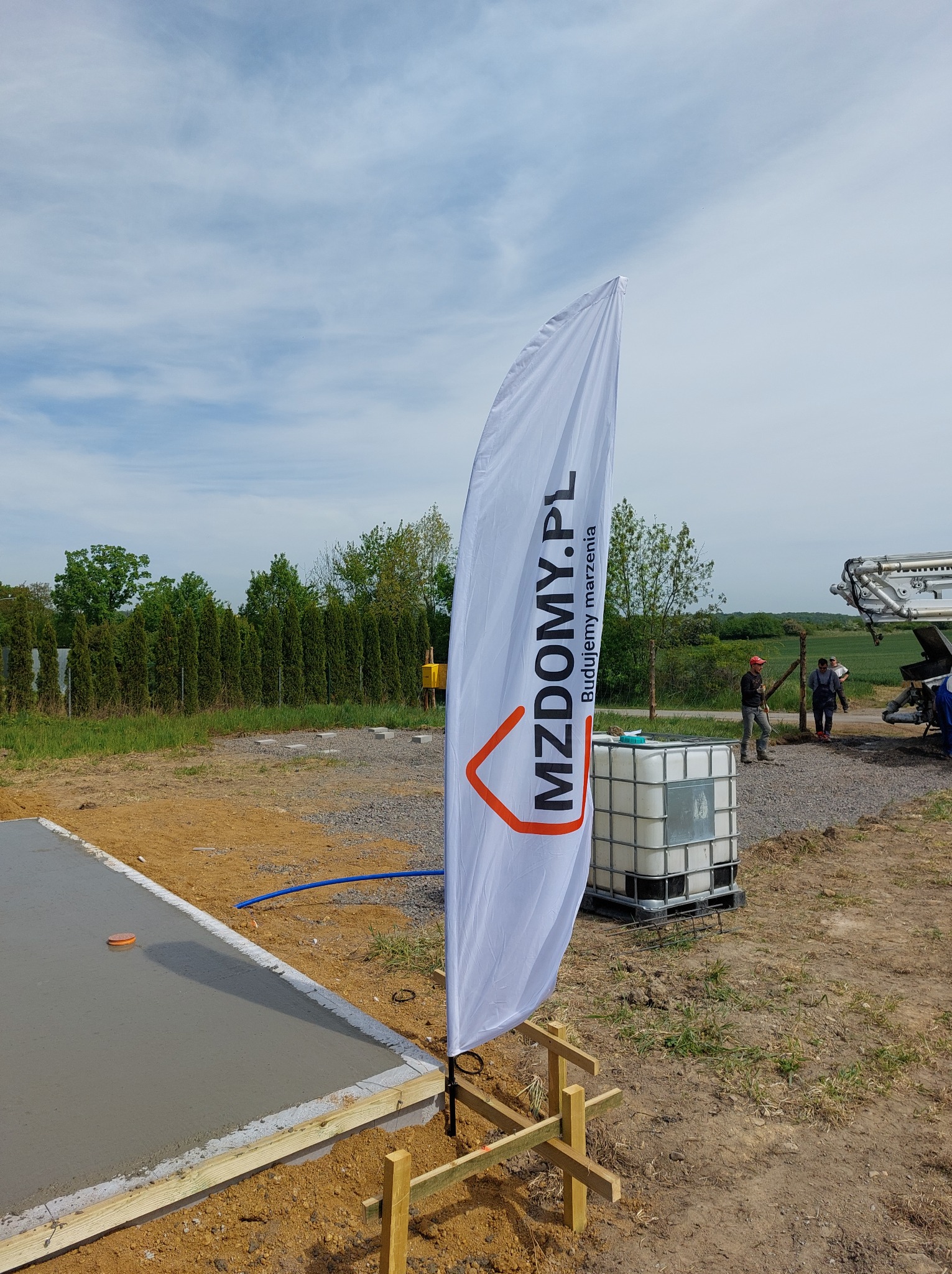 Wylany fundament pod budowę domu, widoczny baner reklamowy firmy budowlanej MZDOMY.PL, w tle pracownicy i pompa do betonu.