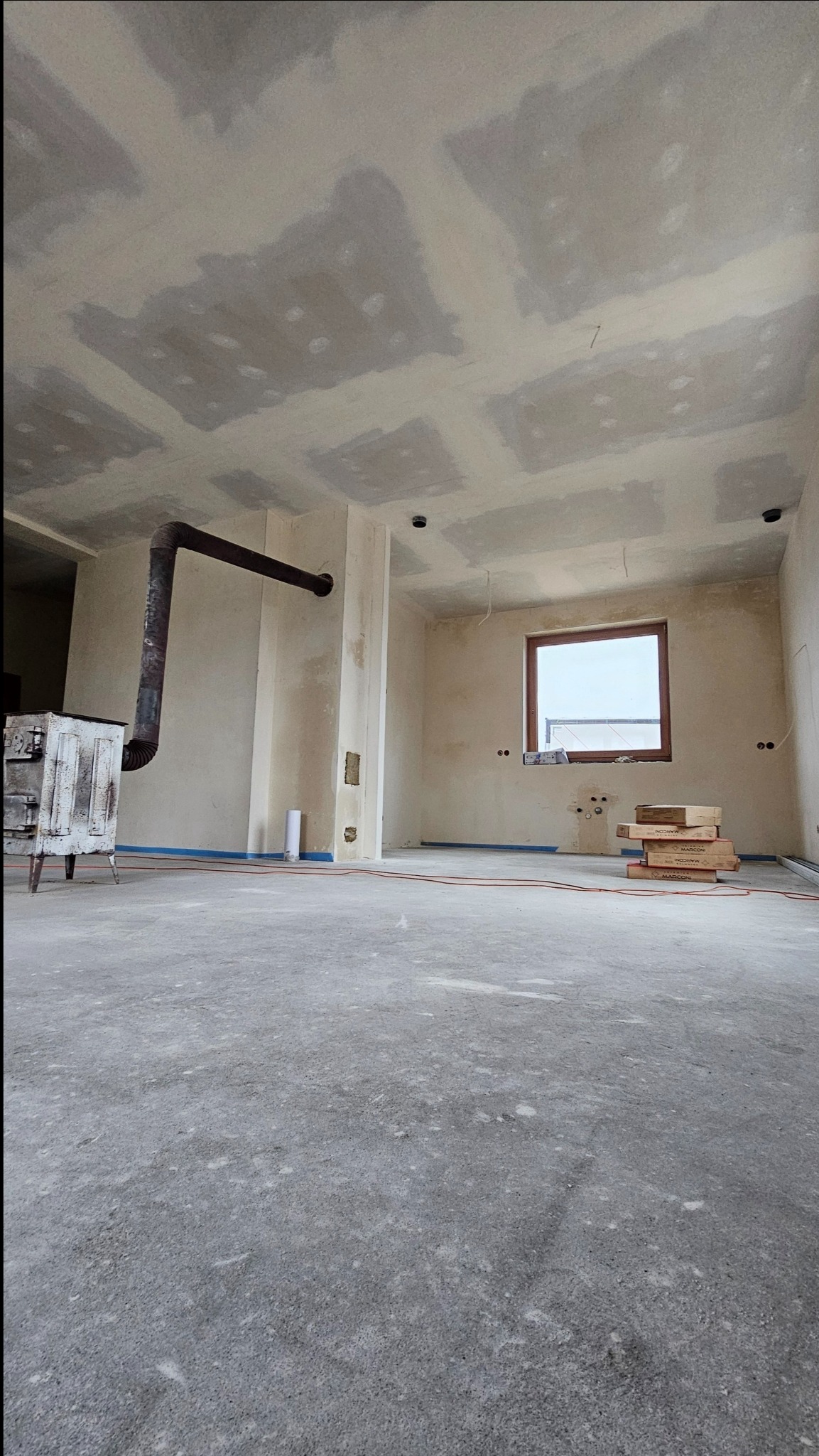 Surowy stan wnętrza domu w trakcie wykańczania: ściany po szpachlowaniu, piec wolnostojący z rurą, okno z widokiem na zewnątrz, podłoga wylewka, puszki elektryczne i przewody na ścianach.