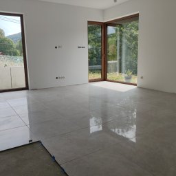 CraftNatura - Wnętrze w trakcie układania jasnoszarych płytek podłogowych, widoczne listwy dystansowe i niezakończona powierzchnia, duże okna z widokiem na zieleń.