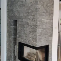 Wykończony narożny kominek z szarej cegły z czarną obudową i drewnem w środku, widoczny fragment podłogi z szarych płytek i lodówki w tle.