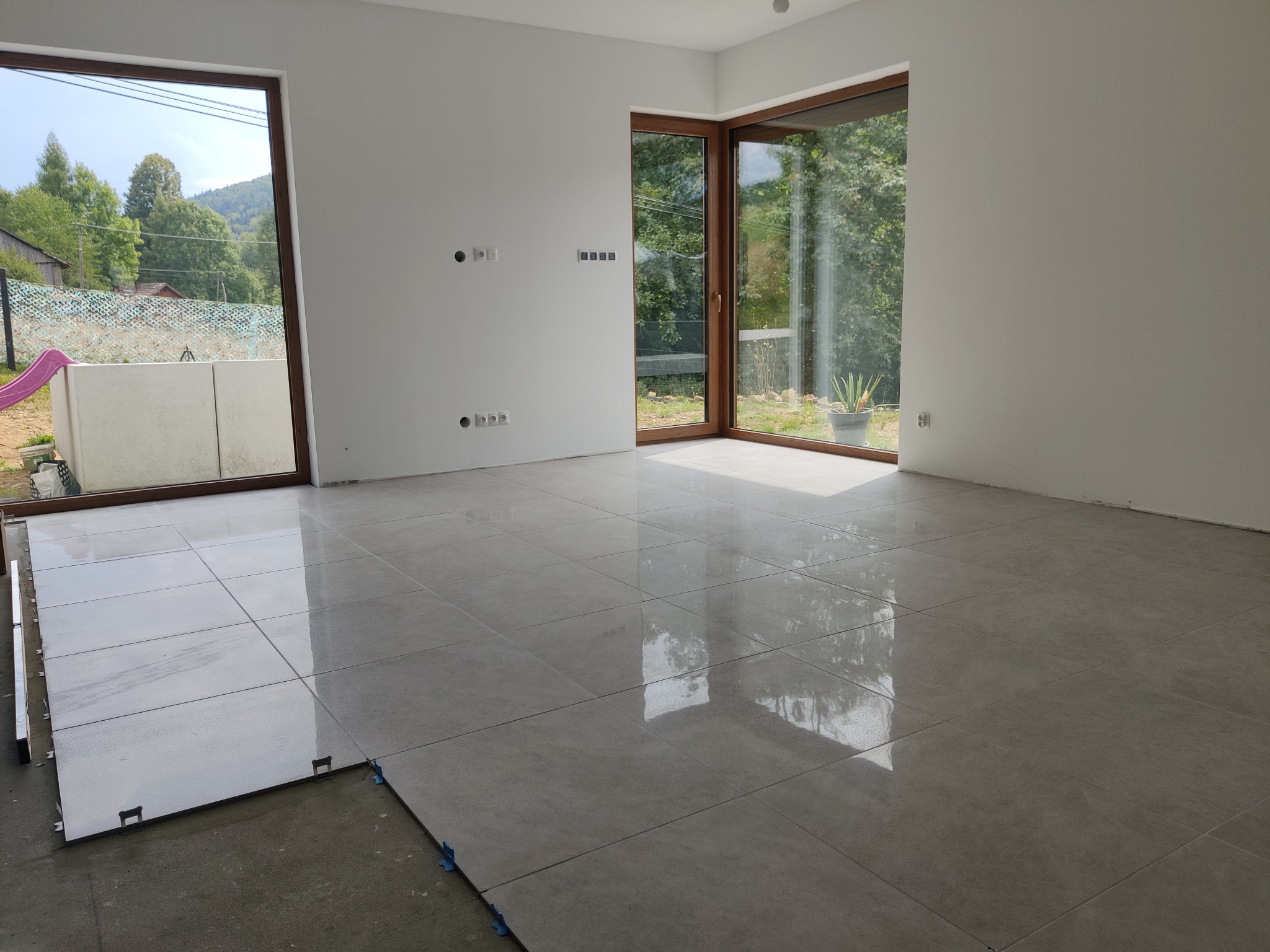 Wnętrze w trakcie układania jasnoszarych płytek podłogowych, widoczne listwy dystansowe i niezakończona powierzchnia, duże okna z widokiem na zieleń.