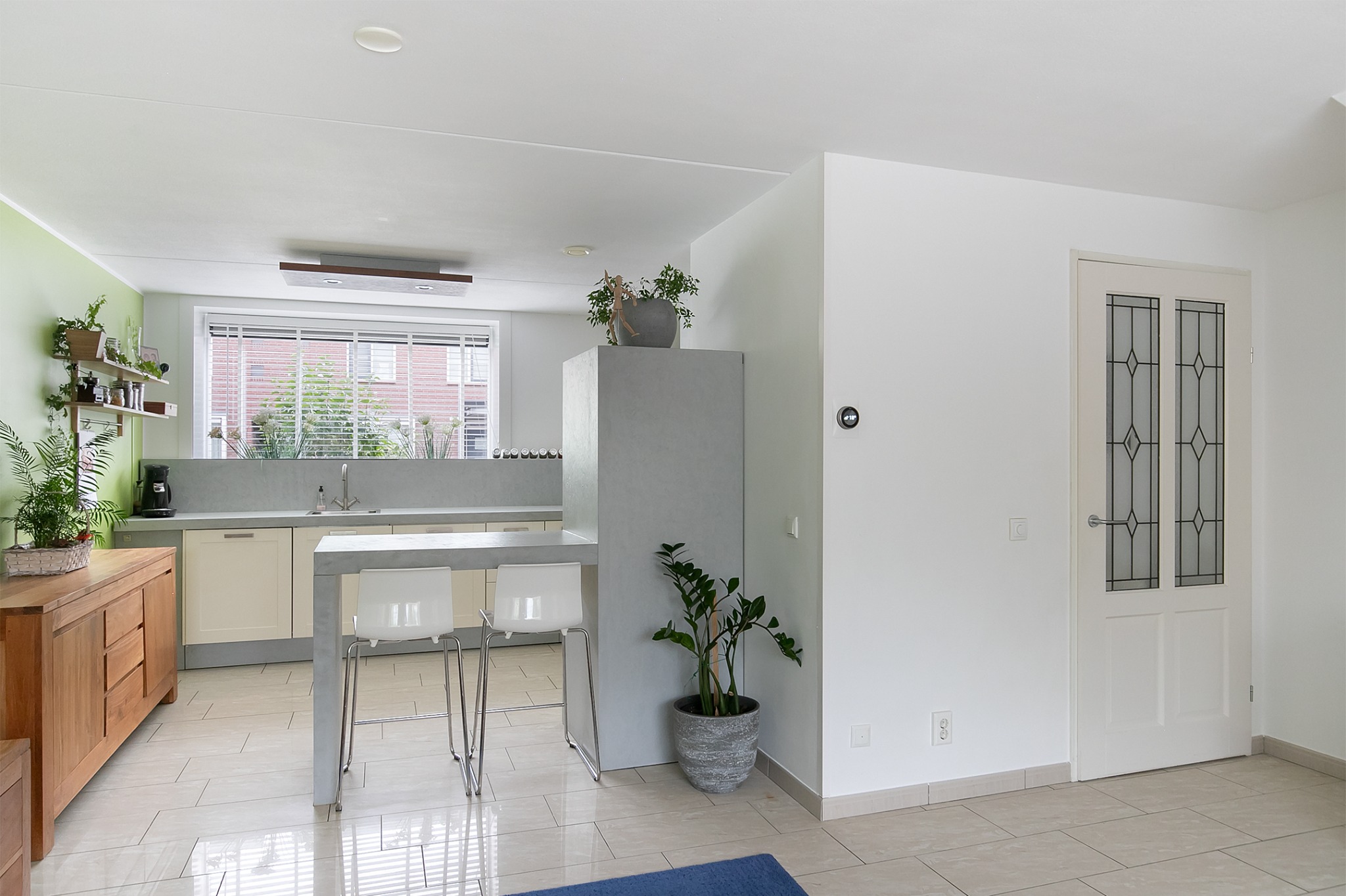 Jasna kuchnia z betonowym blatem, białymi szafkami, drewnianą komodą i roślinami doniczkowymi, widok na przeszklone drzwi.