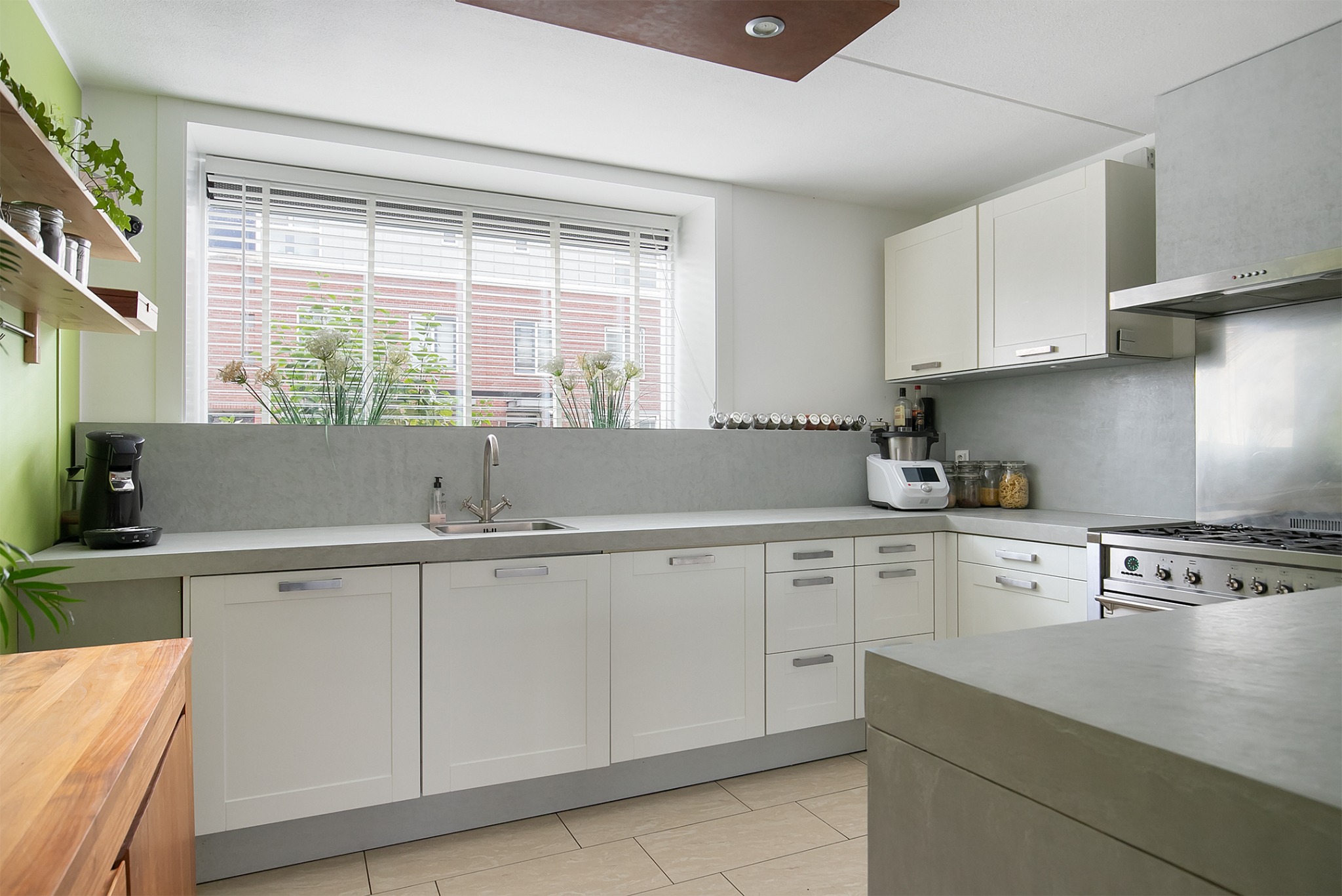 Jasna kuchnia z białymi szafkami, szarym blatem i kuchenką gazową, widok na okno z roślinami i żaluzjami.