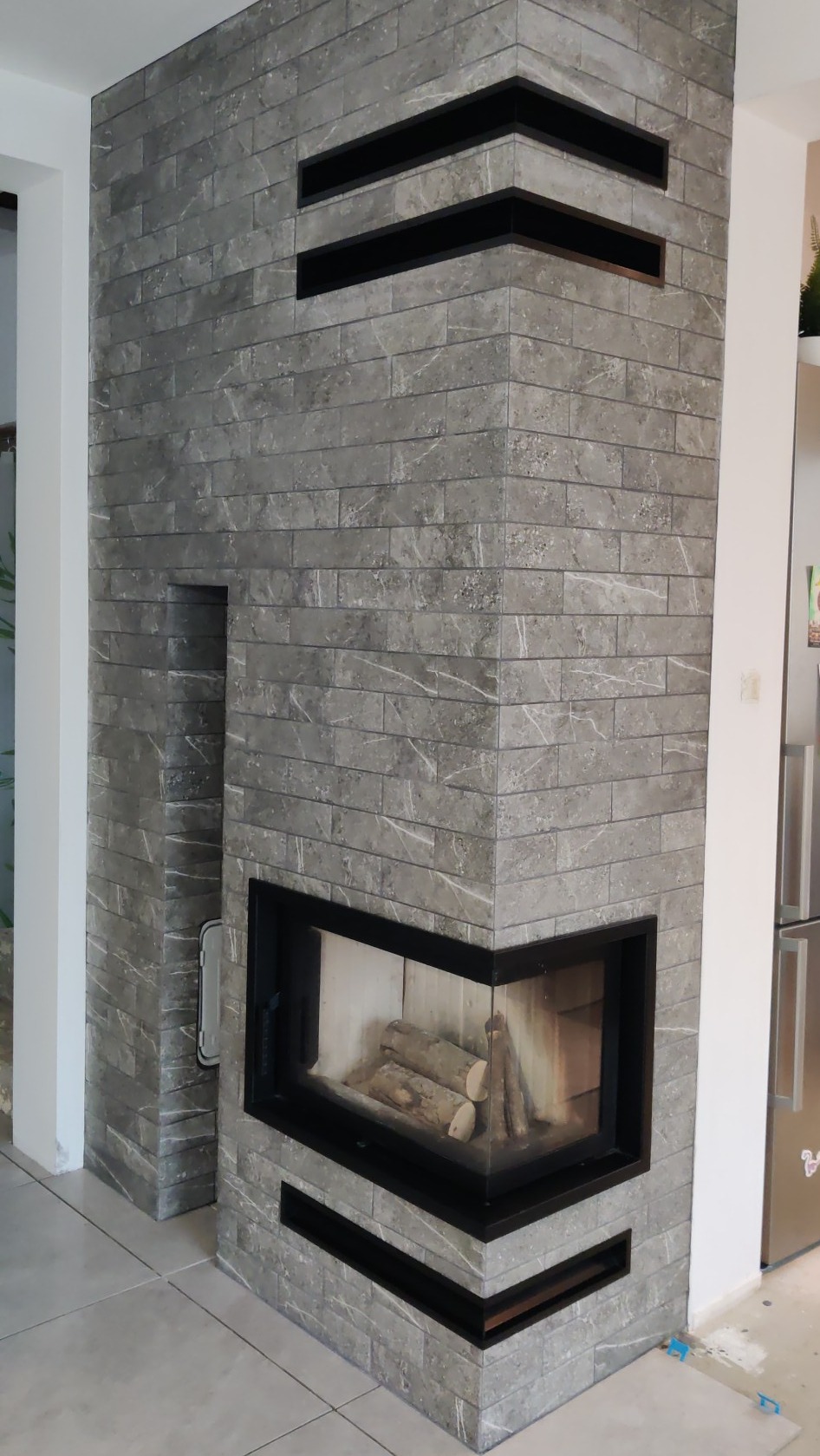 Wykończony narożny kominek z szarej cegły z czarną obudową i drewnem w środku, widoczny fragment podłogi z szarych płytek i lodówki w tle.