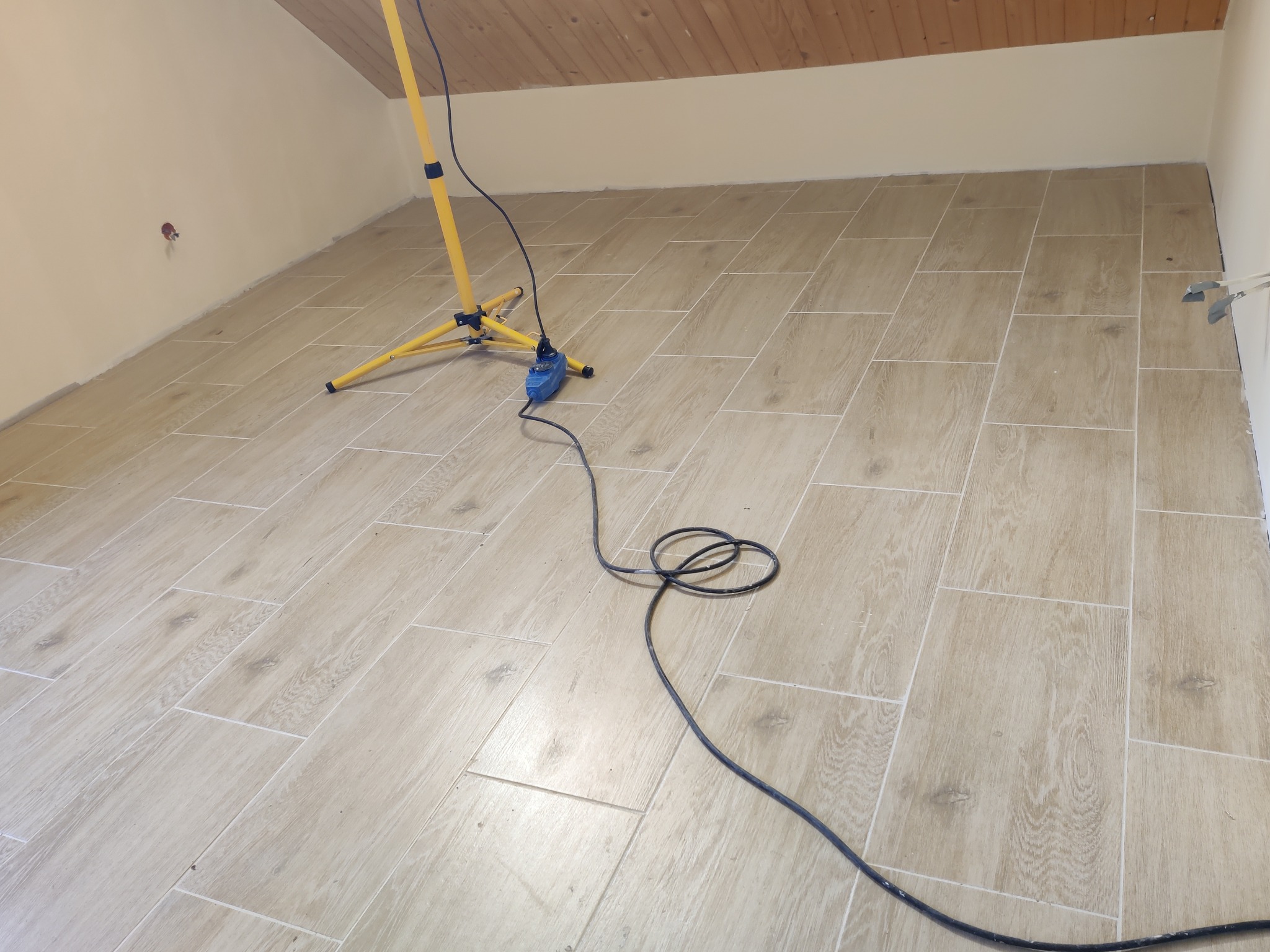 Układanie podłogi z płytek imitujących drewno w pokoju na poddaszu, widoczna lampa budowlana na trójnogu i kabel zasilający.