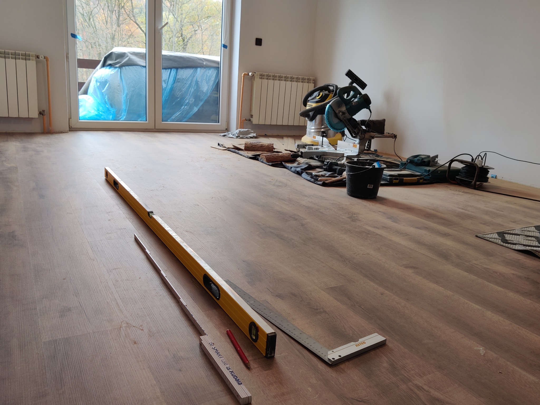 Podłoga z paneli w trakcie układania, widoczny poziomica, kątownik, miara składana i ołówek, w tle piła do paneli i odkurzacz przemysłowy.