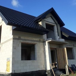 Mateusz Marcinkowski - Dom w trakcie budowy z czarnym dachem i rynnami. Widoczne niezadaszone okna i drzwi. Budynek z białych pustaków, na tle błękitnego nieba i drzew.