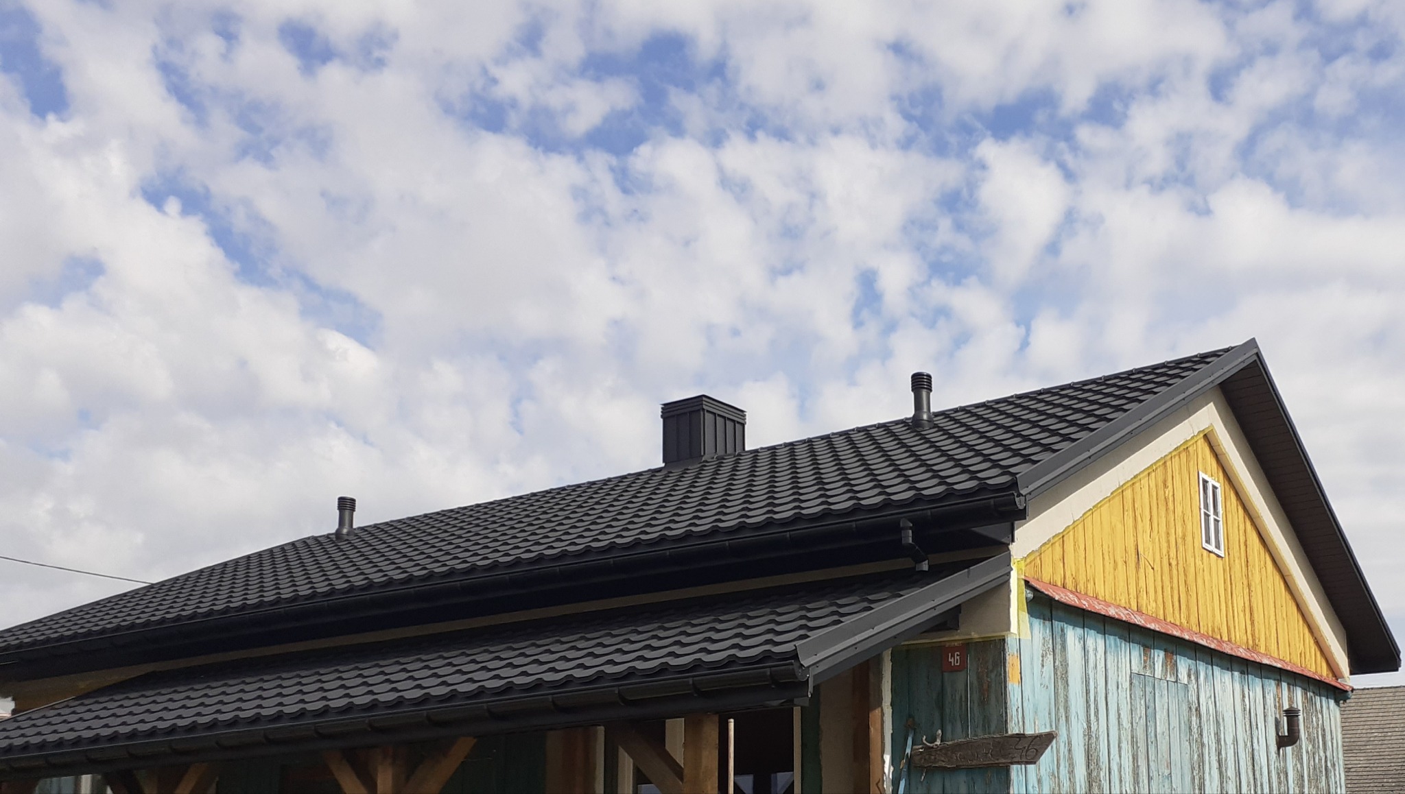 Czarny dach z blachy trapezowej z kominem i rynnami na tle błękitnego nieba z chmurami. Fragment elewacji w kolorach żółtym i niebieskim.