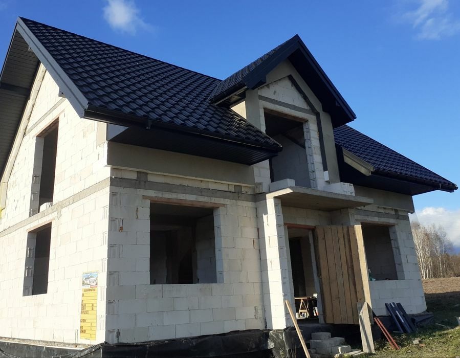 Dom w trakcie budowy z czarnym dachem i rynnami. Widoczne niezadaszone okna i drzwi. Budynek z białych pustaków, na tle błękitnego nieba i drzew.