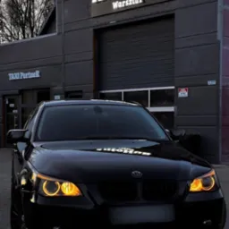 Czarny samochód osobowy przed budynkiem warsztatu samochodowego z widocznym szyldem 'AutoHub Warsztat' i napisem 'TAXI Partner' na sąsiednim wejściu.