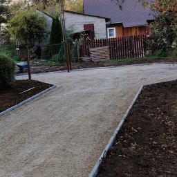 Gospodarstwo rolne Dominik Filipowicz - Przygotowanie podłoża pod drogę dojazdową: ubity tłuczeń ograniczony szarymi obrzeżami, w tle widoczny pracownik z taczką oraz dom jednorodzinny z drewnianym płotem.