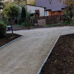 Przygotowanie podłoża pod drogę dojazdową: ubity tłuczeń ograniczony szarymi obrzeżami, w tle widoczny pracownik z taczką oraz dom jednorodzinny z drewnianym płotem.