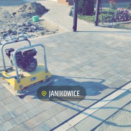 Gospodarstwo rolne Dominik Filipowicz - Żółta zagęszczarka spalinowa na świeżo ułożonej, szarej kostce brukowej przed domem z numerem 26, widok na częściowo wykonany chodnik i przygotowany teren pod dalsze prace brukarskie. W tle zielony...