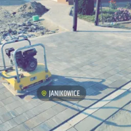 Żółta zagęszczarka spalinowa na świeżo ułożonej, szarej kostce brukowej przed domem z numerem 26, widok na częściowo wykonany chodnik i przygotowany teren pod dalsze prace brukarskie. W tle zielony...