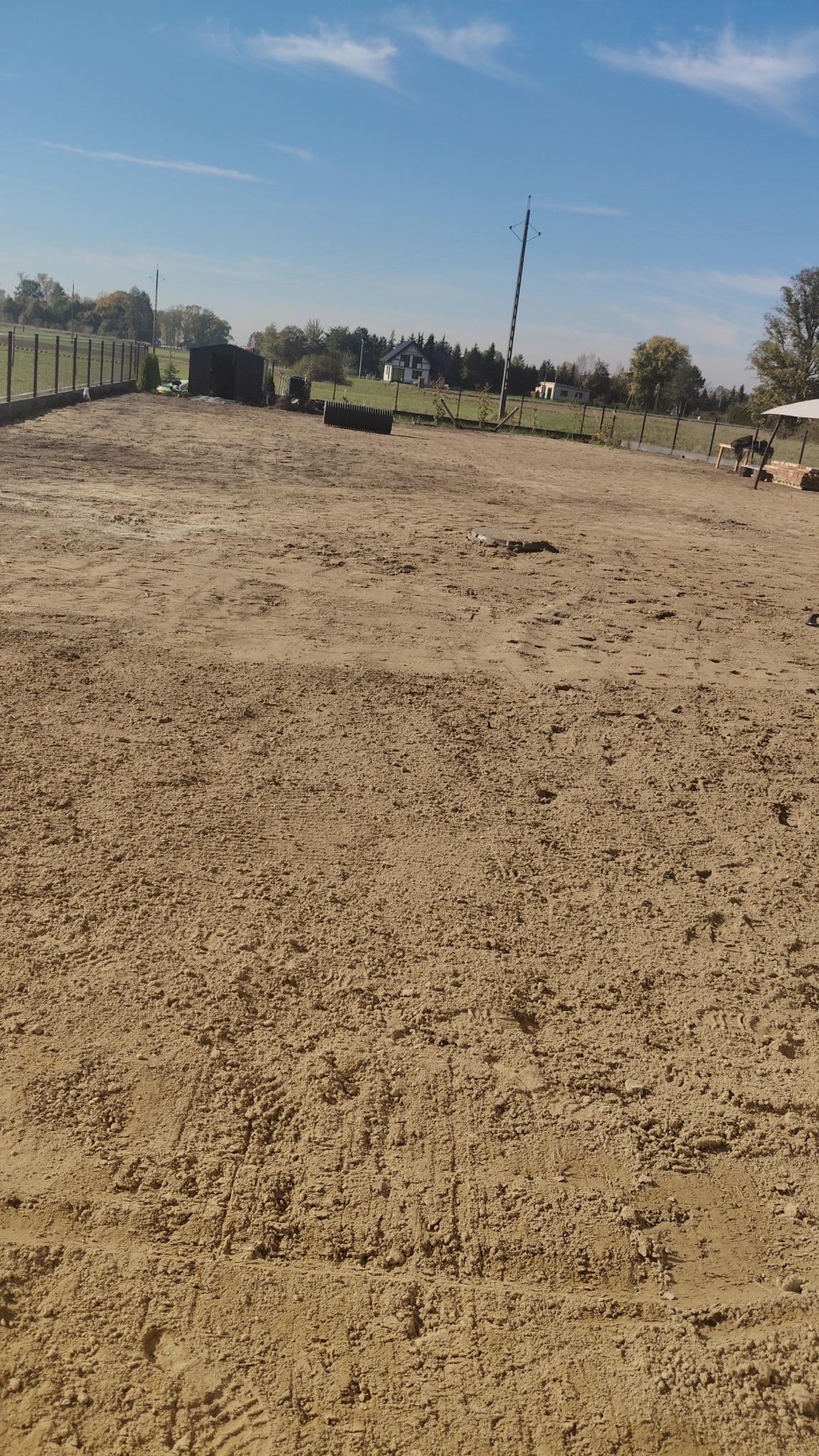 Przygotowany teren pod budowę z widocznymi śladami opon, ogrodzeniem, małym budynkiem gospodarczym i słupem energetycznym w tle pod błękitnym niebem.