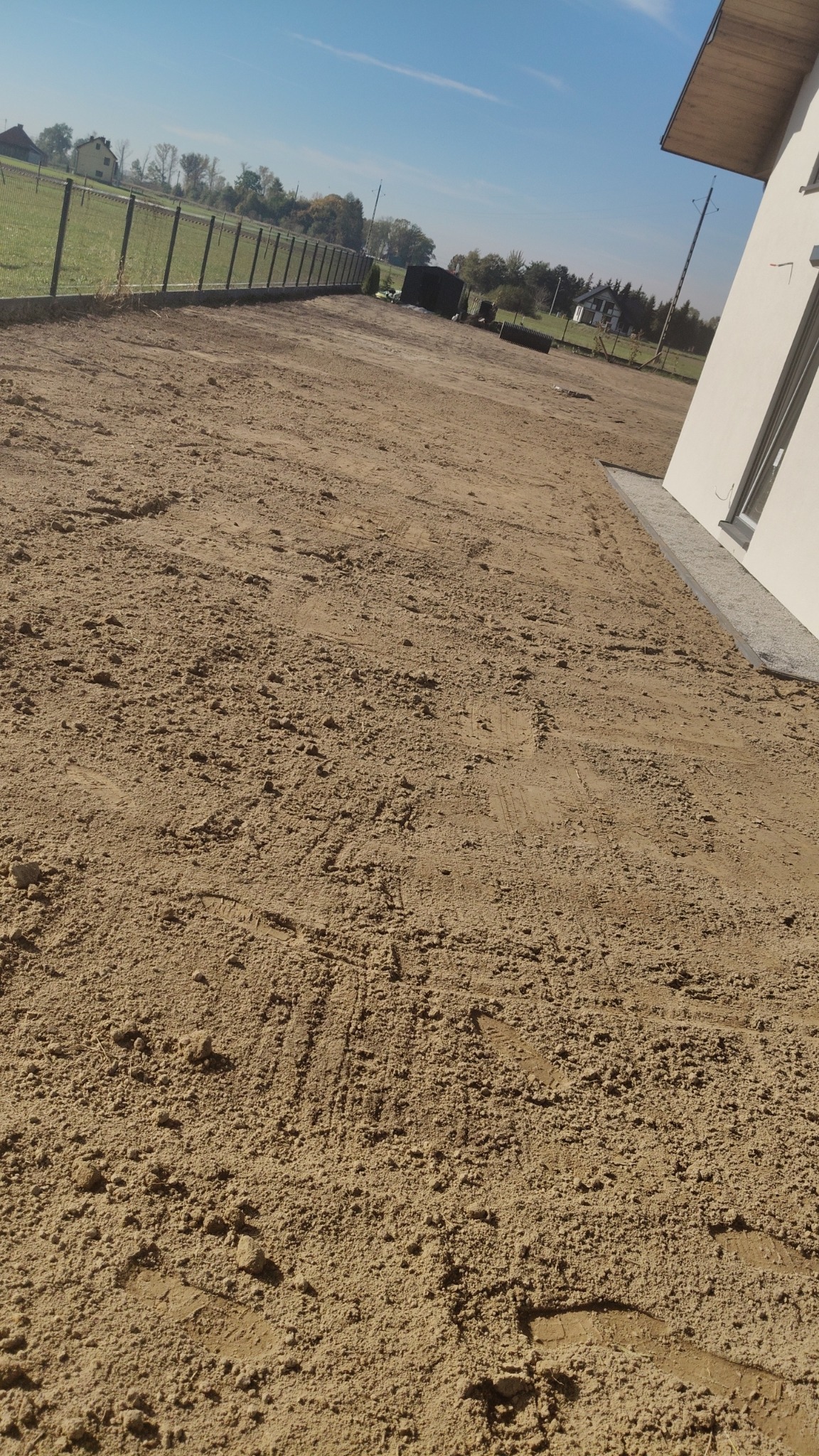 Przygotowany teren pod budowę, wyrównany piasek z widocznymi śladami opon i butów, ogrodzenie z siatki w tle, fragment budynku z oknem po prawej stronie.