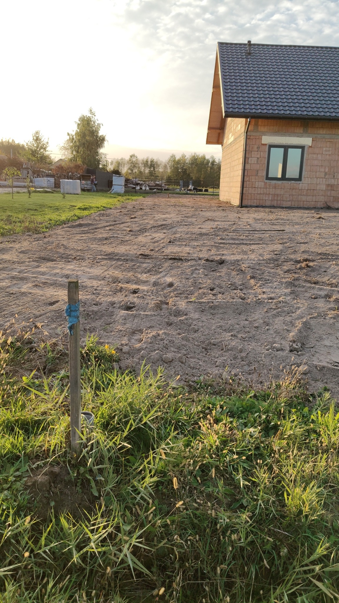 Przygotowany teren pod budowę domu jednorodzinnego, widoczna świeżo wyrównana ziemia, fragment budynku z cegły silikatowej i drewniany palik z niebieską taśmą oznaczający punkt pomiarowy.