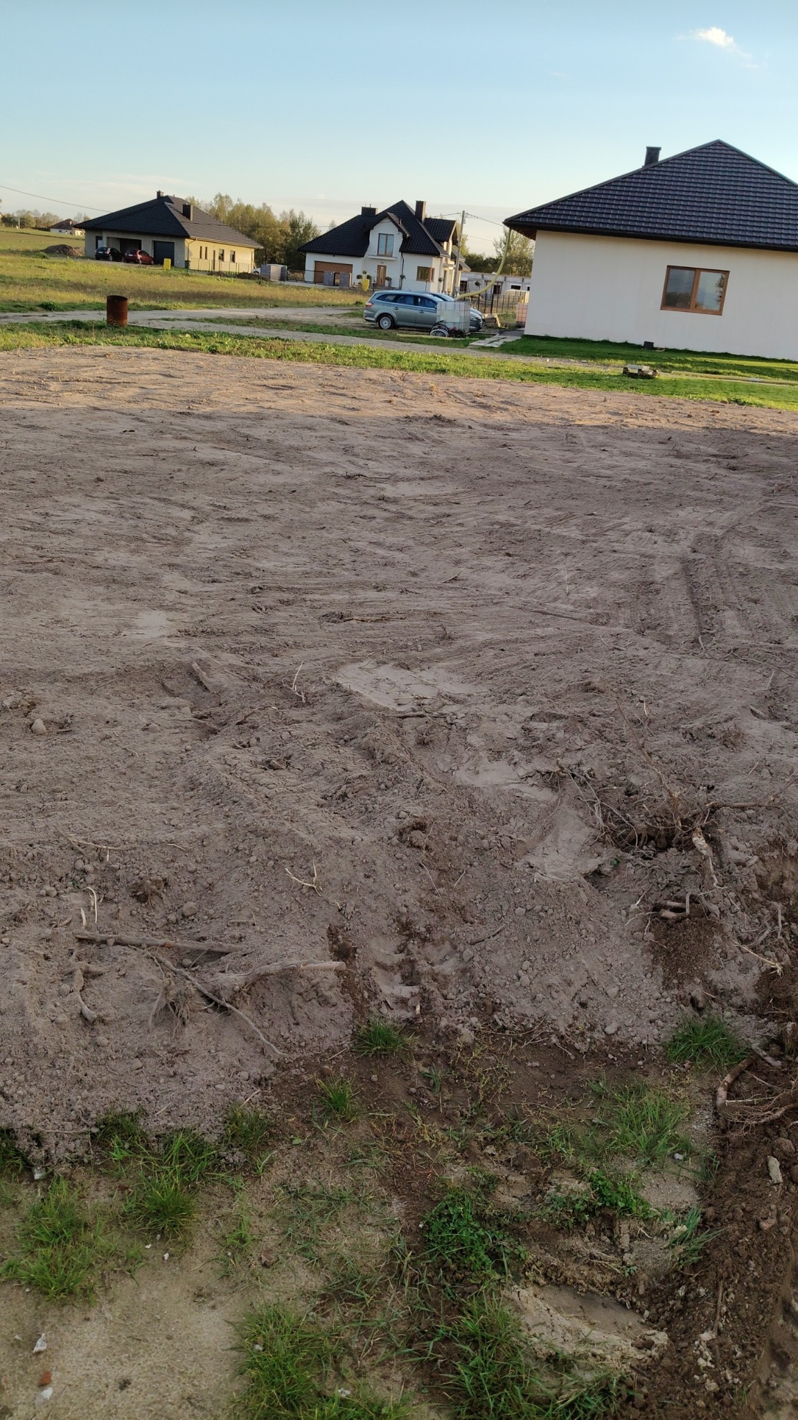 Przygotowany teren pod budowę domu jednorodzinnego, widoczne ślady opon, w tle zabudowa mieszkalna i błękitne niebo.