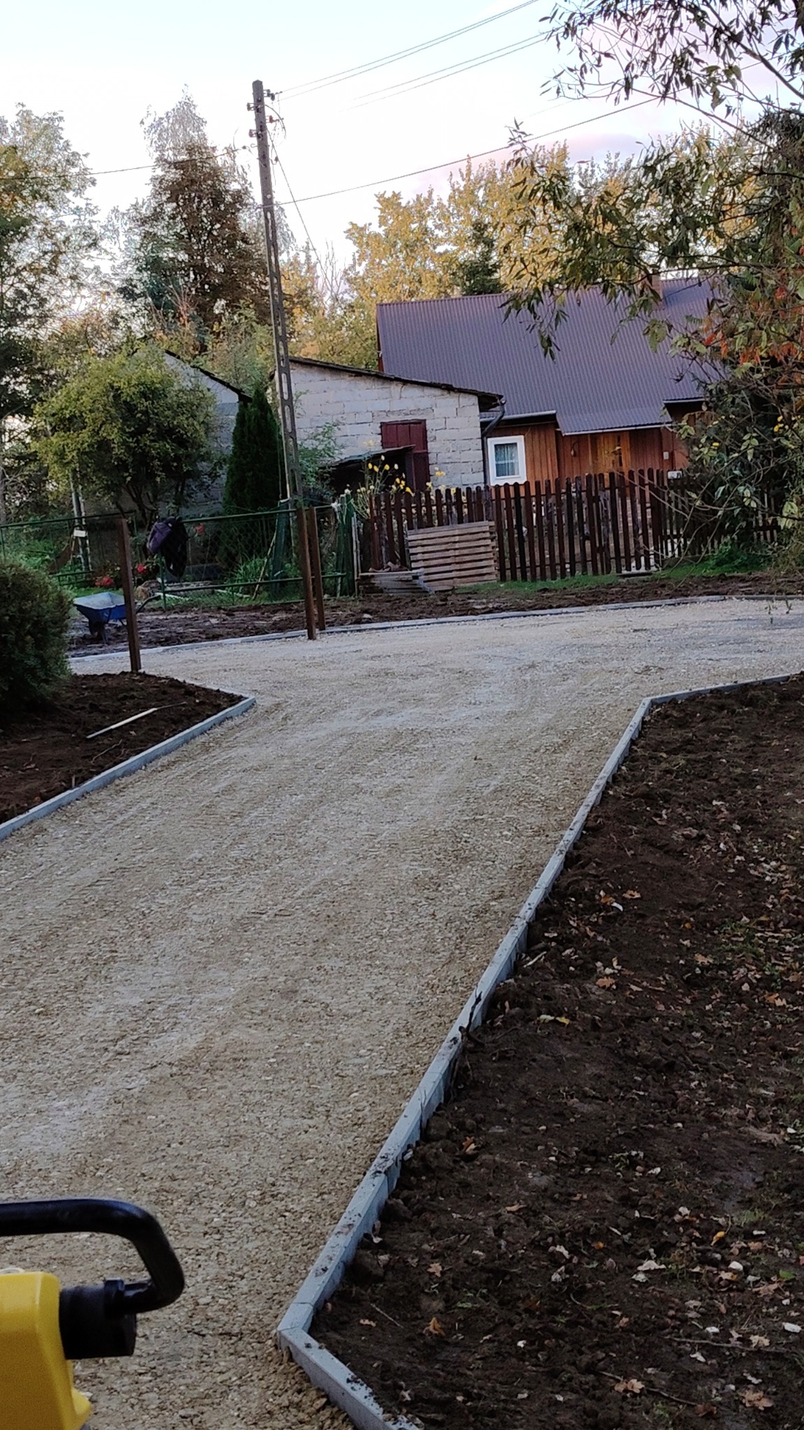 Przygotowanie podłoża pod drogę dojazdową: ubity tłuczeń ograniczony szarymi obrzeżami, w tle widoczny pracownik z taczką oraz dom jednorodzinny z drewnianym płotem.