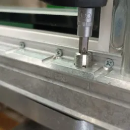 Wkręcanie śruby w metalowy element okna narzędziem z nasadką, widoczne oznaczenia na profilu okiennym.