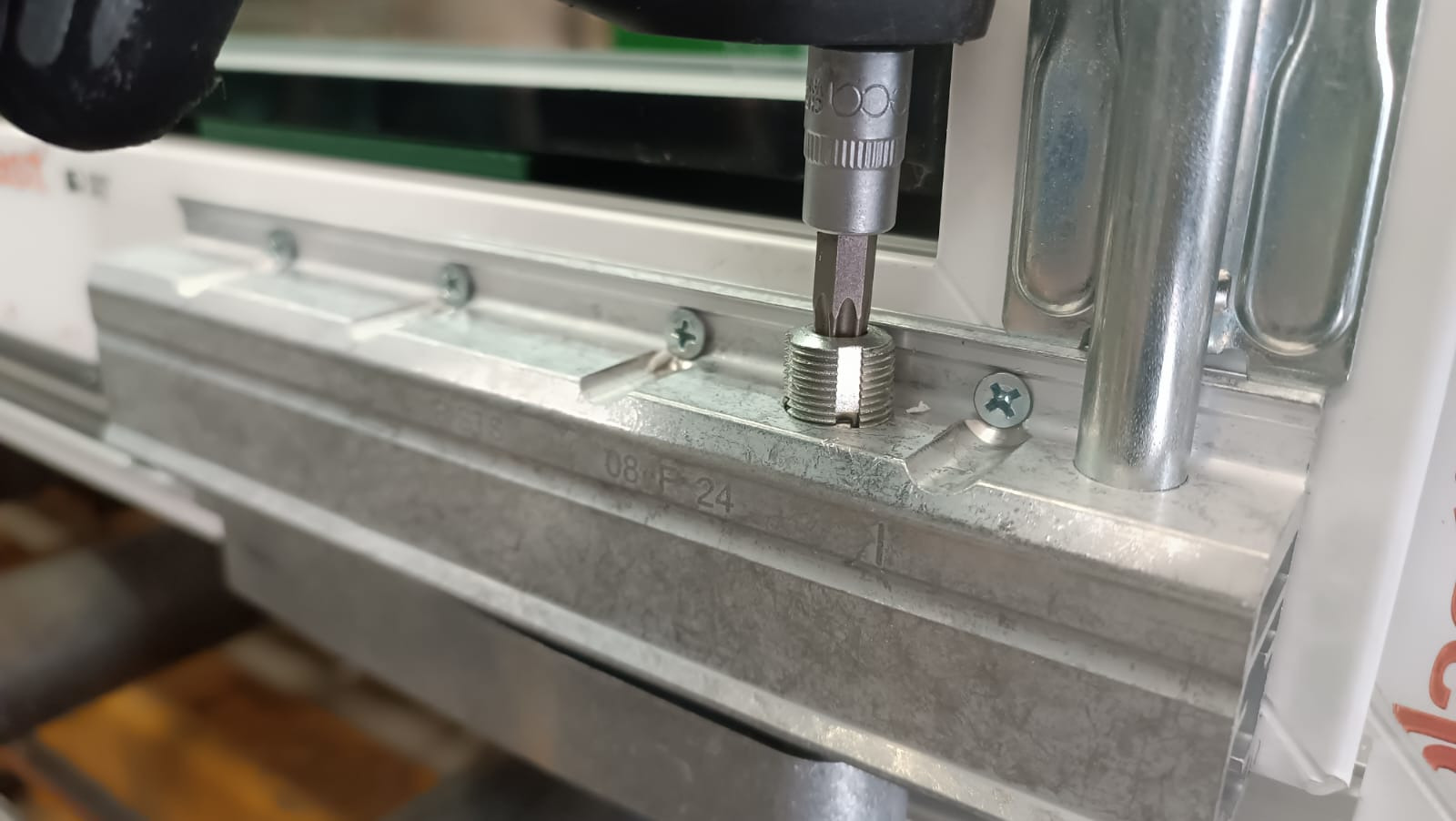 Wkręcanie śruby w metalowy element okna narzędziem z nasadką, widoczne oznaczenia na profilu okiennym.