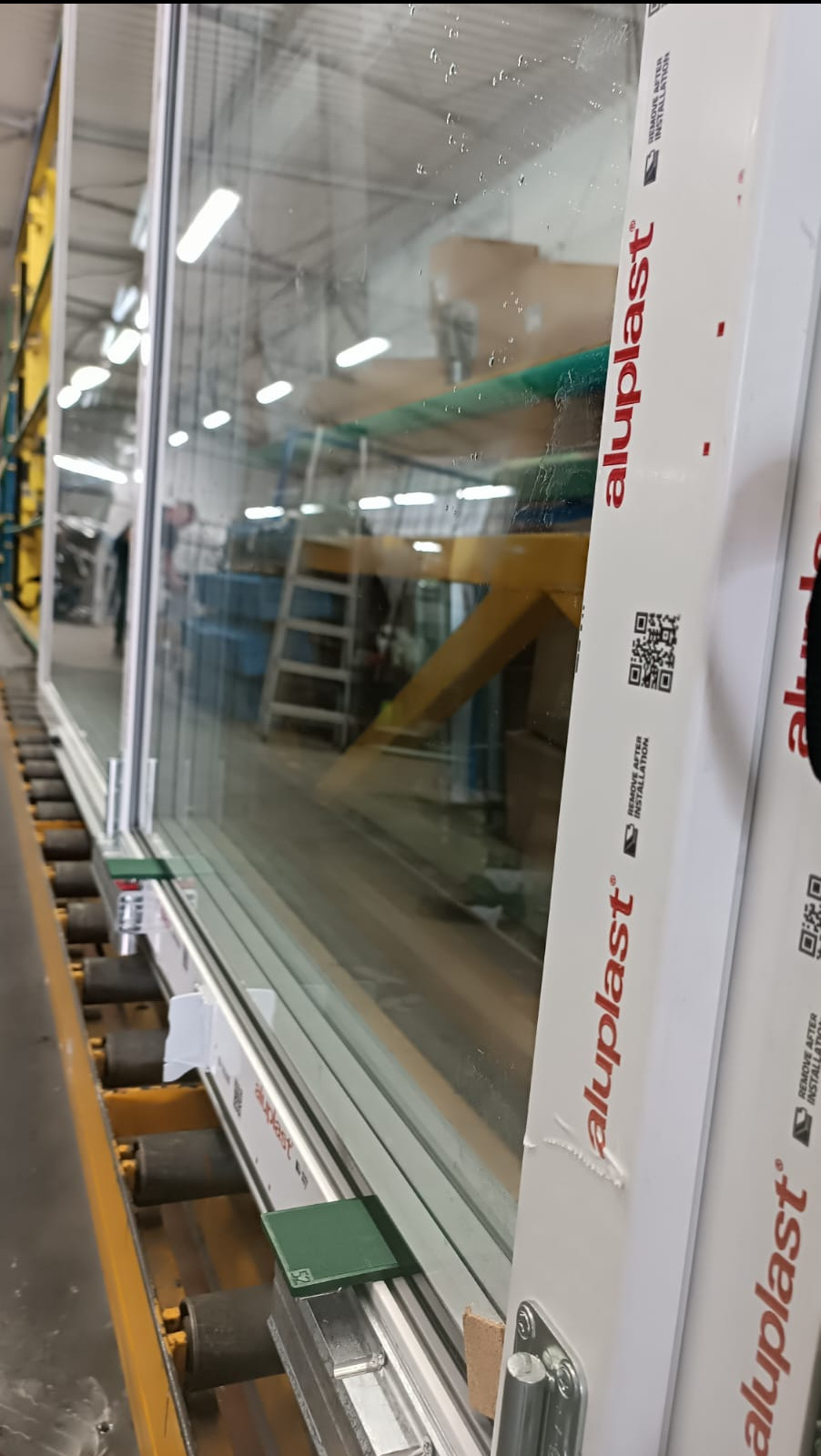 Nowe okno z szybą zespoloną na linii produkcyjnej, widoczny profil Aluplast z kodem QR i oznaczeniem 'Remove After Installation', w tle hala fabryczna.