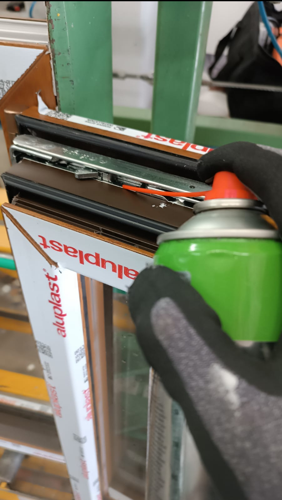 Smarowanie mechanizmu okna PCV Aluplast zielonym sprayem przez osobę w czarnych rękawicach roboczych.