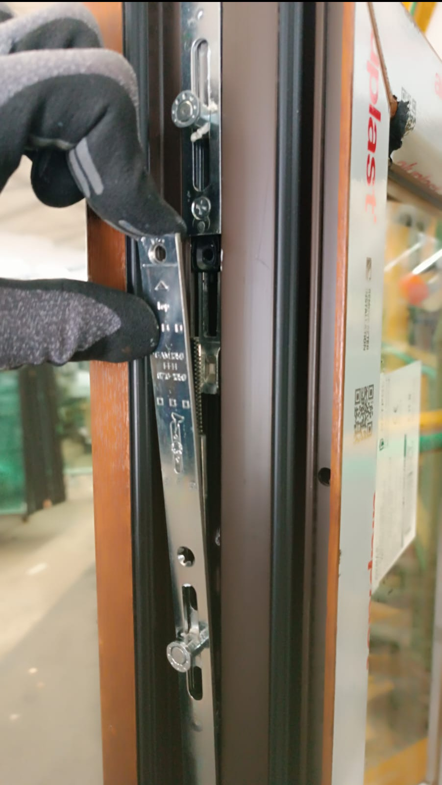 Ręka w roboczej rękawicy reguluje metalowy mechanizm zamka w brązowej ramie okiennej, widoczne śruby i oznaczenia producenta na listwie zamka.