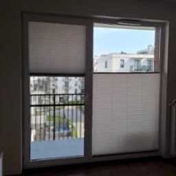 Zasłonięte okno balkonowe z zamontowanymi, częściowo opuszczonymi, białymi osłonami okiennymi typu 'góra-dół', przez które widać zabudowę miejską.
