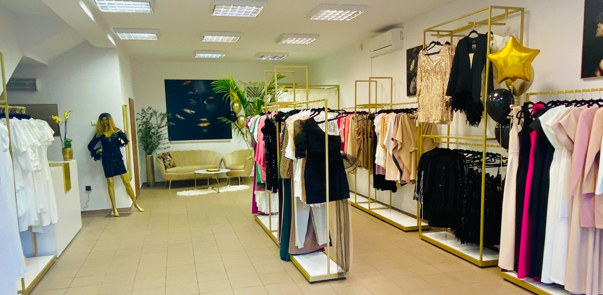 Wnętrze butiku odzieżowego z ubraniami na złotych wieszakach, manekin w czarnej sukience, sofa i obraz na ścianie.