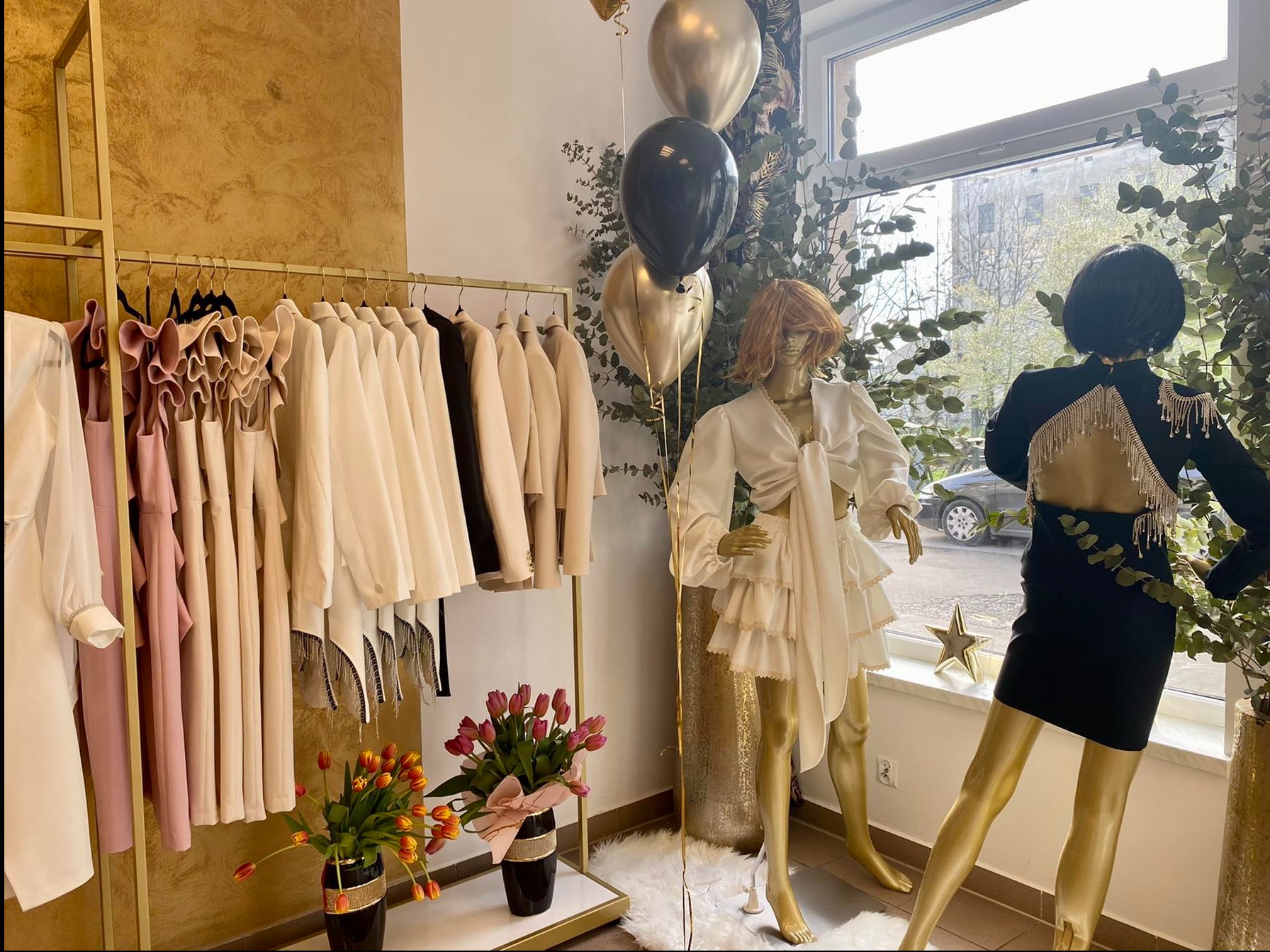 Wnętrze butiku z ubraniami na złotym wieszaku, manekiny w kreacjach, bukiety tulipanów w czarnych wazonach, złote i czarne balony, dekoracja z eukaliptusa przy oknie.