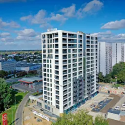 Wysoki budynek mieszkalny z balkonami, widok z góry na miasto w tle, częściowo jeszcze w trakcie budowy.