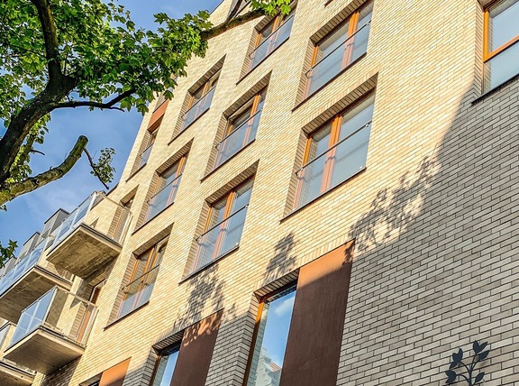 Fasada budynku z jasnej cegły z oknami o drewnianych ramach i szklanymi balkonami. Na ścianie cień rzucany przez drzewo. W tle fragment błękitnego nieba.