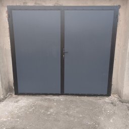 TOM-STAL TOMASZ KŁAKULAK - Szara, dwuskrzydłowa brama garażowa z czarną ramą, osadzona w surowym, betonowym otworze. Widoczne niedoskonałości tynku i szorstka posadzka.