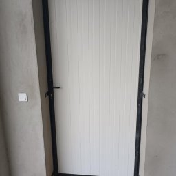 TOM-STAL TOMASZ KŁAKULAK - Biała brama garażowa z czarną, metalową ramą, zamontowana w surowym, betonowym otworze. Widoczny fragment ściany z włącznikiem światła.