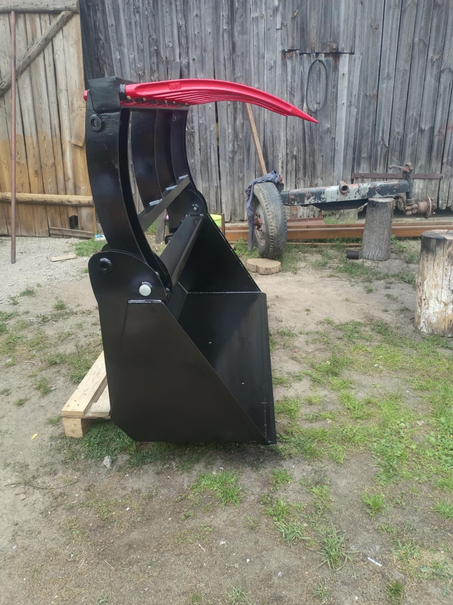 Czarny, metalowy chwytak do ładowarki z czerwonymi elementami, stojący na palecie przed drewnianym budynkiem i fragmentem podwozia pojazdu.