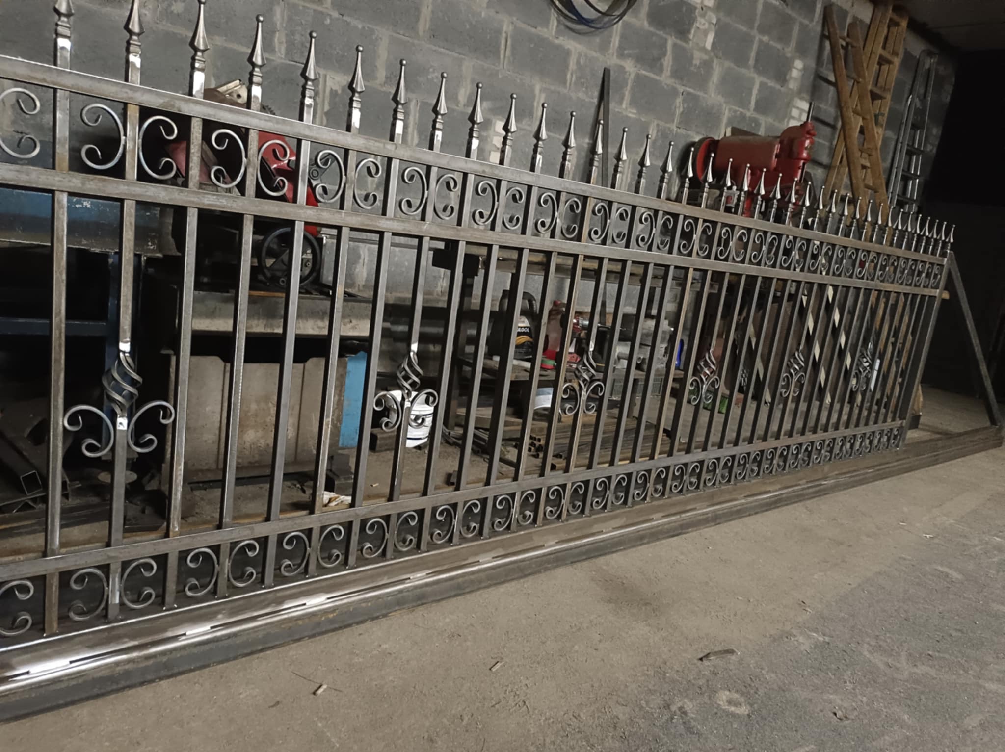 Surowa, metalowa brama z dekoracyjnymi elementami w warsztacie. Widoczne narzędzia i ceglana ściana w tle. Brama w trakcie produkcji, przed malowaniem.