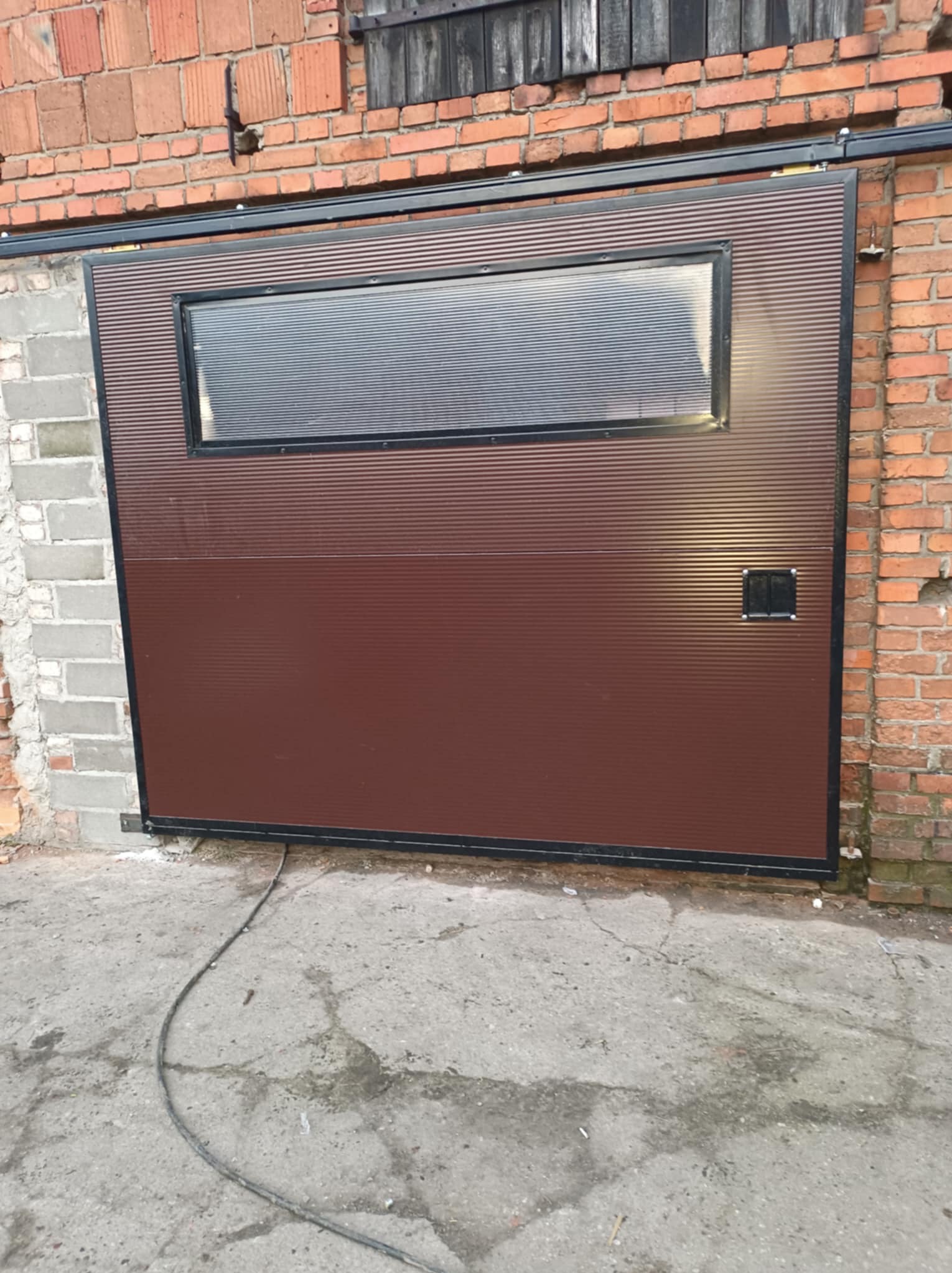 Brązowa brama przesuwna z poziomymi przetłoczeniami i oknem z pleksi, zamontowana na tle ceglanego budynku. Widoczny betonowy podjazd i kabel zasilający.