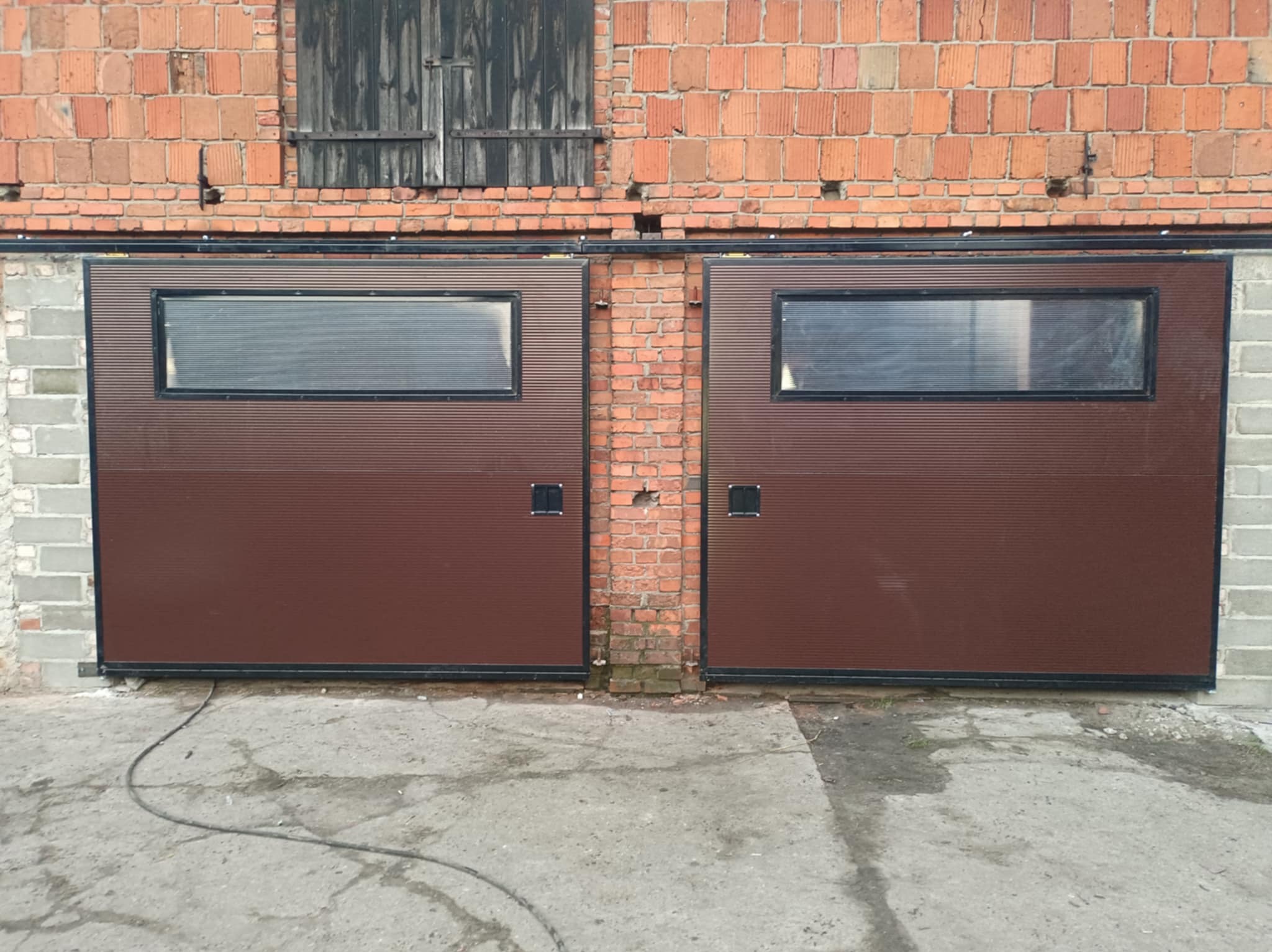 Dwie brązowe bramy garażowe segmentowe z oknami, zamontowane w ceglanym budynku. Widoczne detale konstrukcyjne i faktura materiałów. Ujęcie z bliska, symetryczne.
