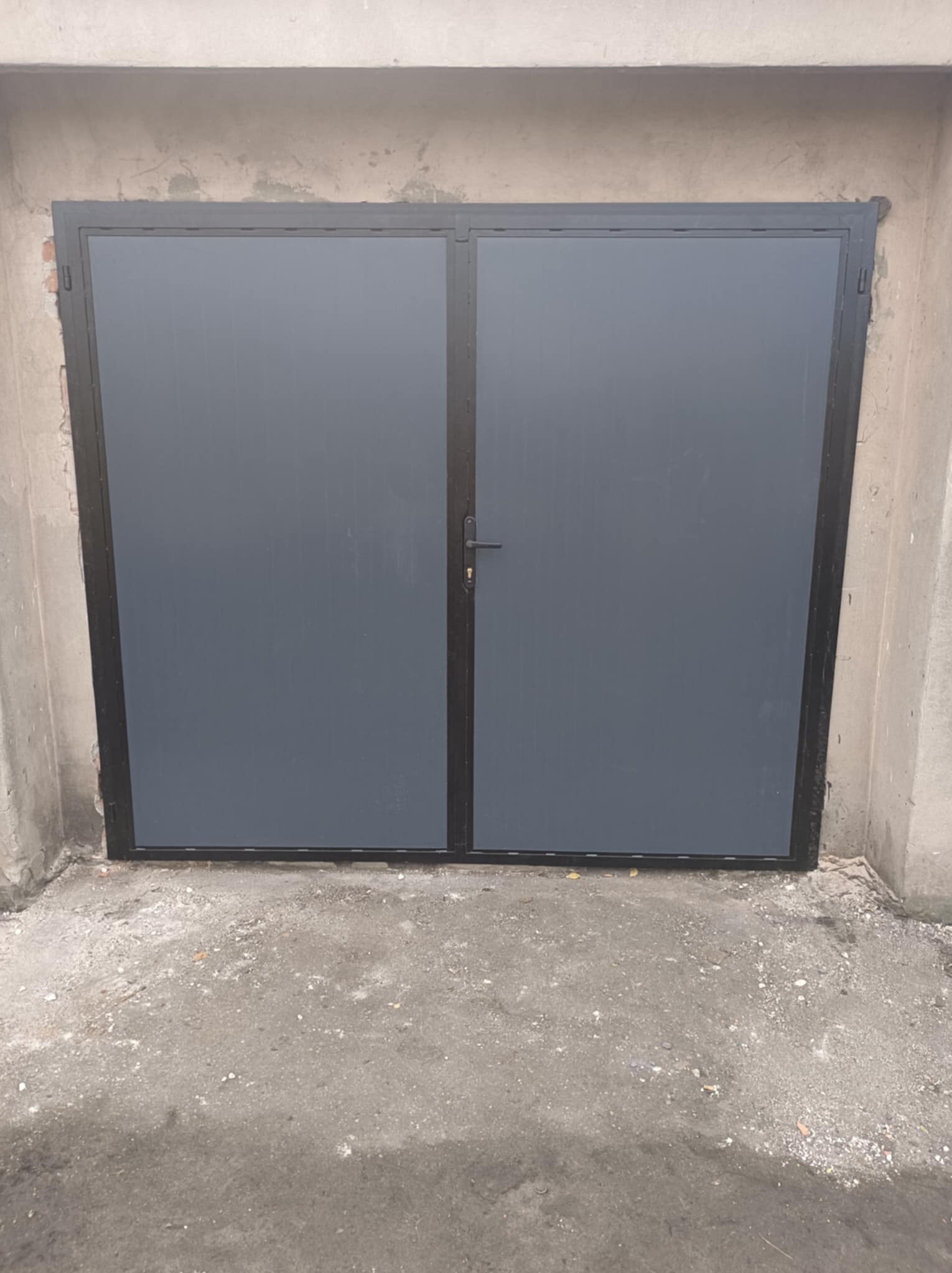 Szara, dwuskrzydłowa brama garażowa z czarną ramą, osadzona w surowym, betonowym otworze. Widoczne niedoskonałości tynku i szorstka posadzka.