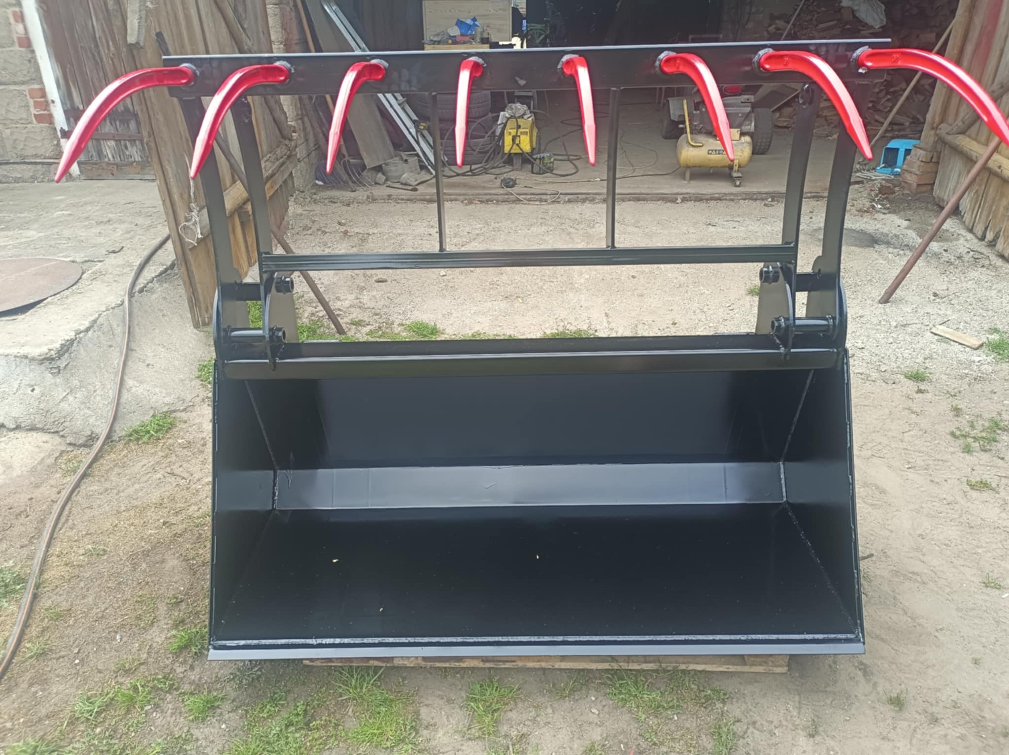 Czarny, metalowy kosz z czerwonymi uchwytami, prawdopodobnie do traktora, wykonany na zamówienie. Widoczne ślady spawania i świeża farba.