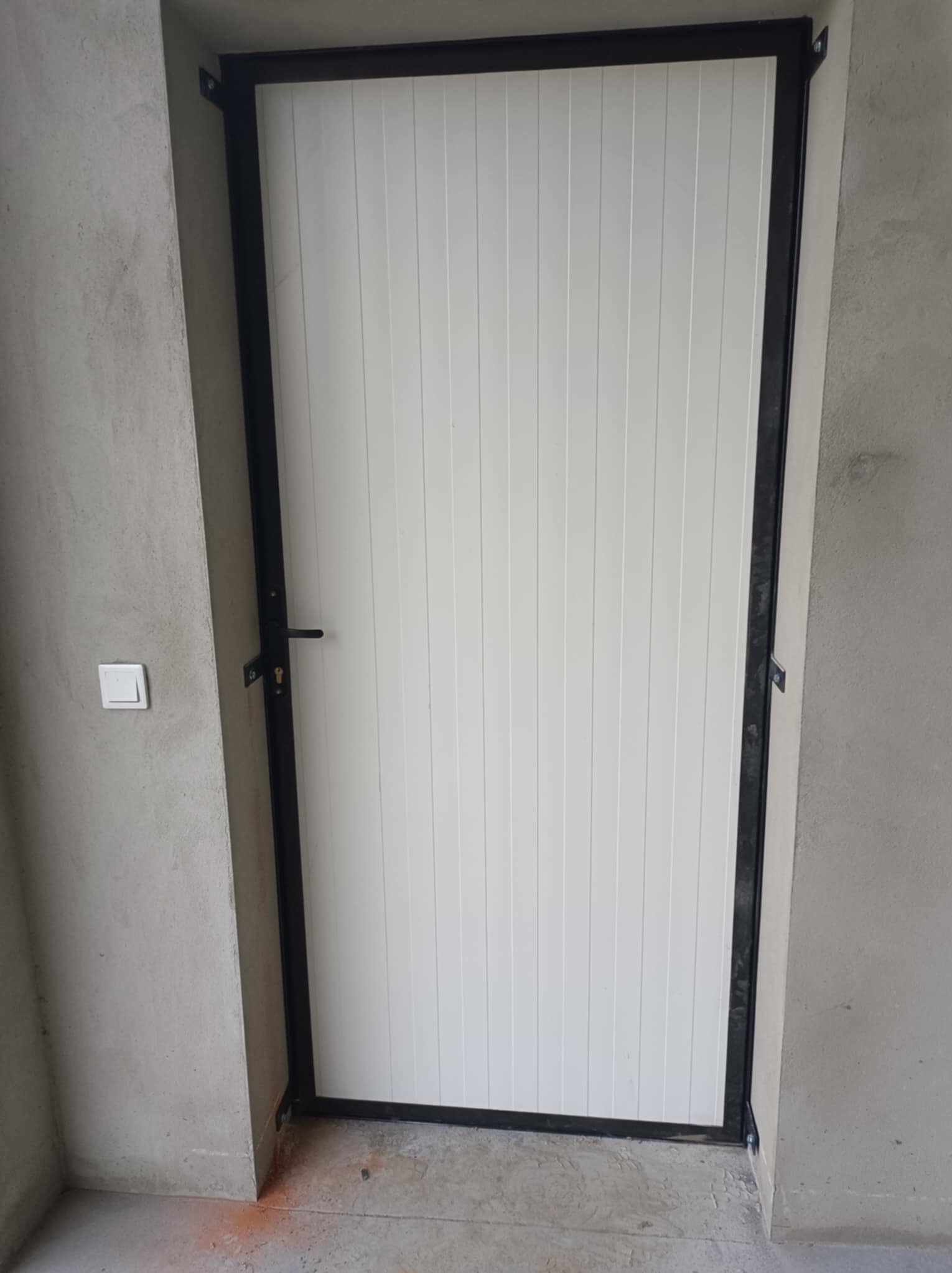 Biała brama garażowa z czarną, metalową ramą, zamontowana w surowym, betonowym otworze. Widoczny fragment ściany z włącznikiem światła.