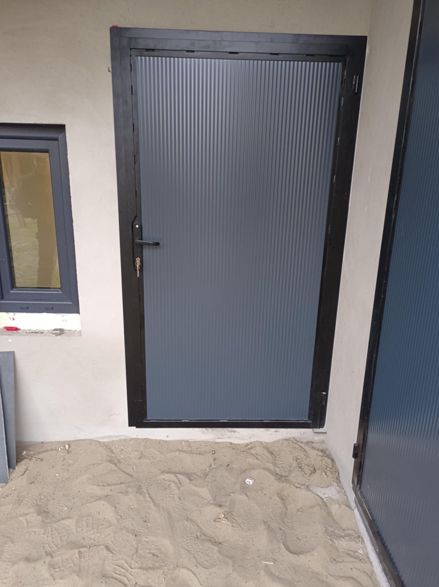 Szara, metalowa furtka z kluczem, zamontowana w surowym otoczeniu. Widoczny fragment okna i piasek na podłożu. Furtka z wypełnieniem z blachy trapezowej.