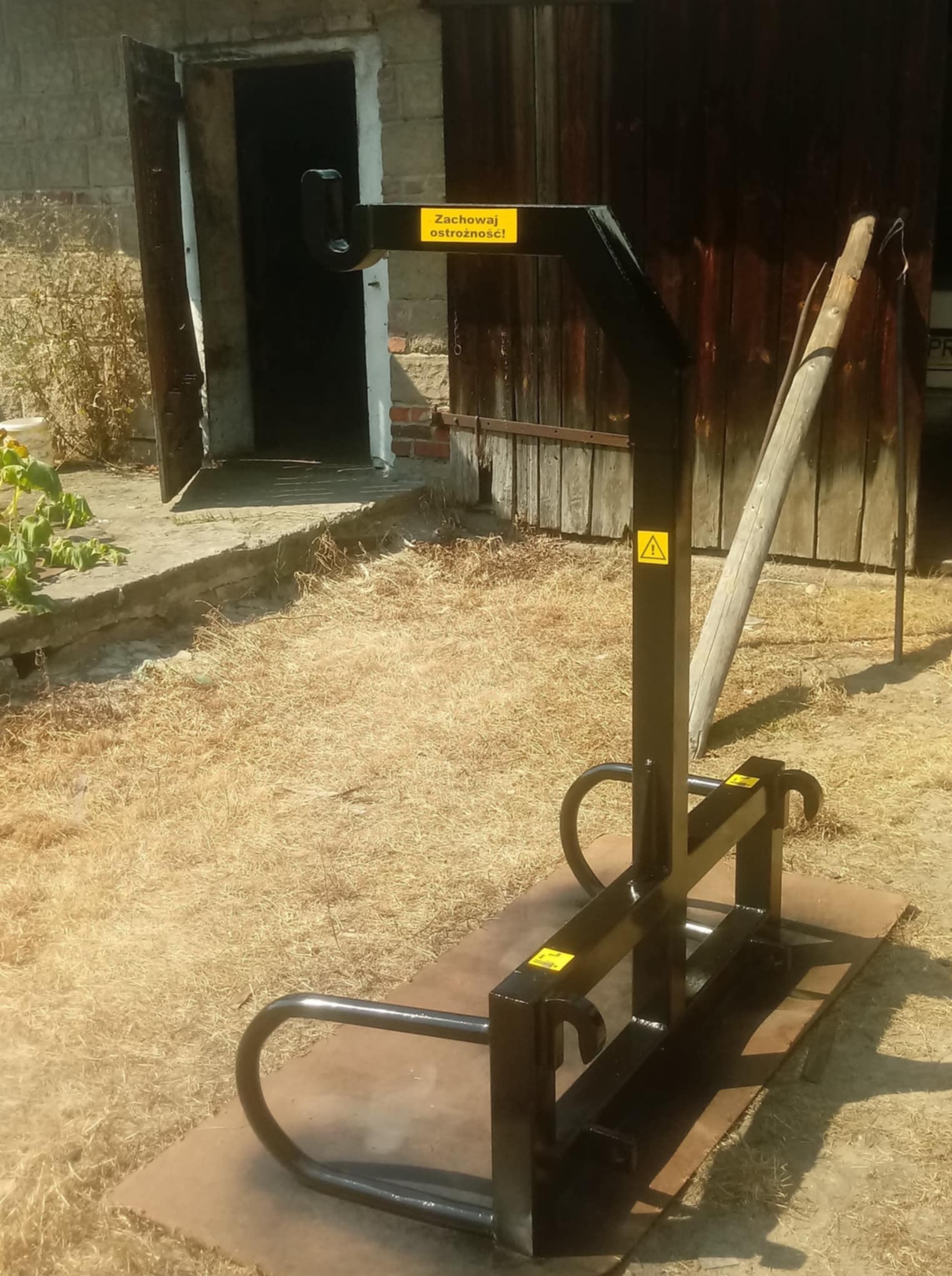 Czarna, metalowa konstrukcja z żółtymi naklejkami ostrzegawczymi na zewnątrz, na tle budynku gospodarczego z otwartymi drzwiami i drewnianą ścianą.
