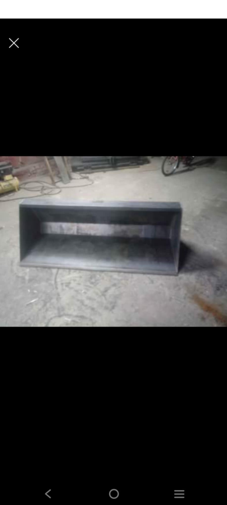 Metalowa skrzynia o prostokątnym kształcie, wykonana z blachy, widoczna na betonowej posadzce w warsztacie. W tle narzędzia i rower.