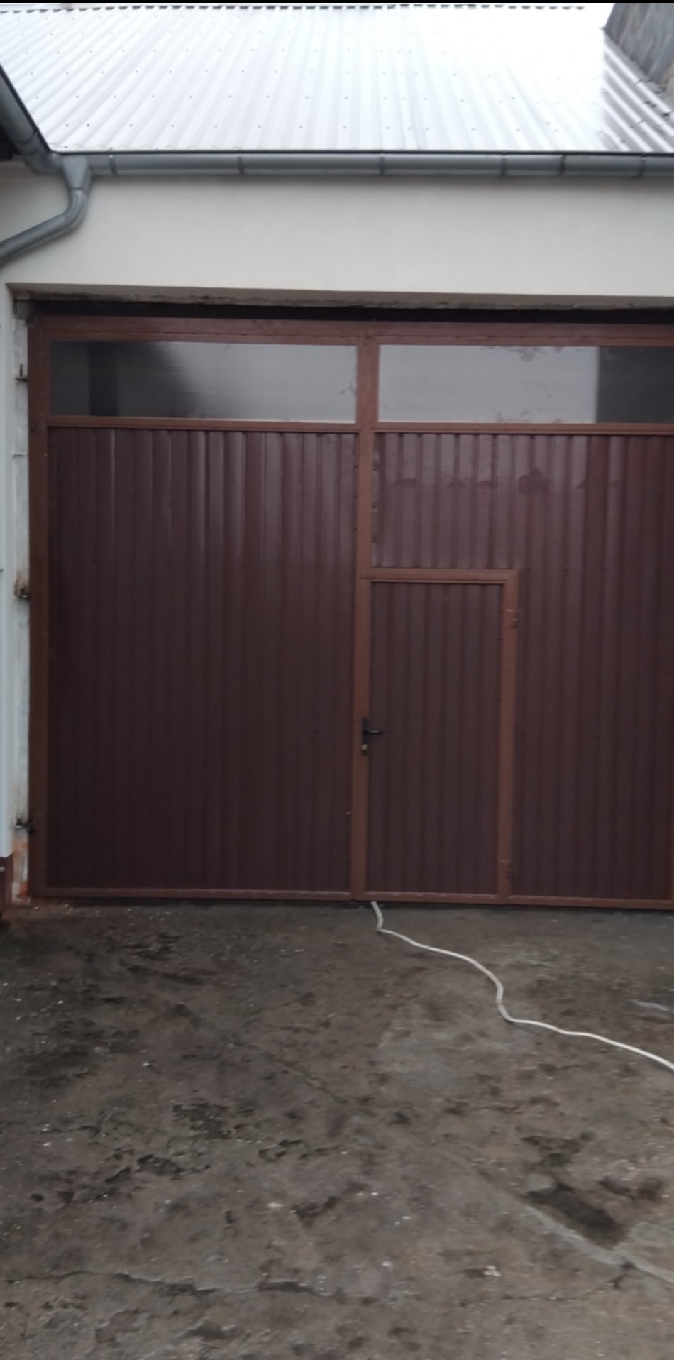 Brązowa brama garażowa z oknem i małym wejściem, wykonana z blachy falistej. Widoczny zamek i kabel na posadzce. Ujęcie z bliska, na zewnątrz budynku.