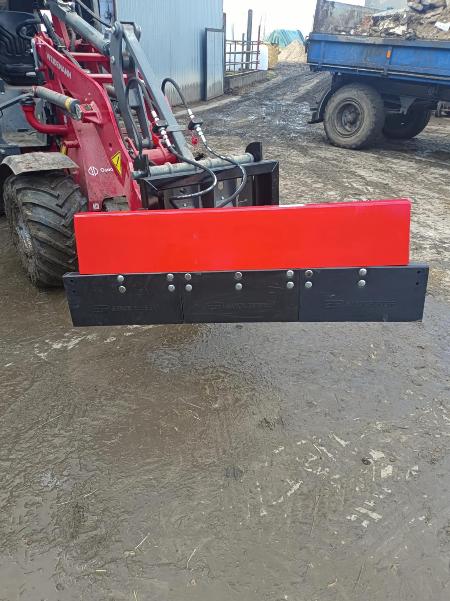 Czerwony ładowacz Weidemann z zamontowaną czarną gumową listwą zgarniającą Sanok Rubber, stojący na błotnistym podłożu. W tle przyczepa i budynek gospodarczy.