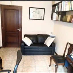 Gabinet w Gdańsku z czarną skórzaną sofą, białymi poduszkami, drewnianym biurkiem, krzesłem i półką z książkami, oświetlony ciepłym światłem.