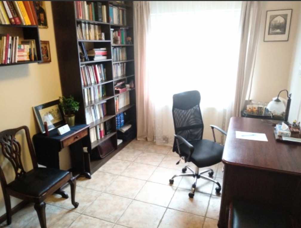 Jasny gabinet z biurkiem, krzesłem obrotowym i regałem pełnym książek, dodatkowo małe biurko z ozdobnym krzesłem, neutralne kolory ścian, okno z zasłonami w tle.